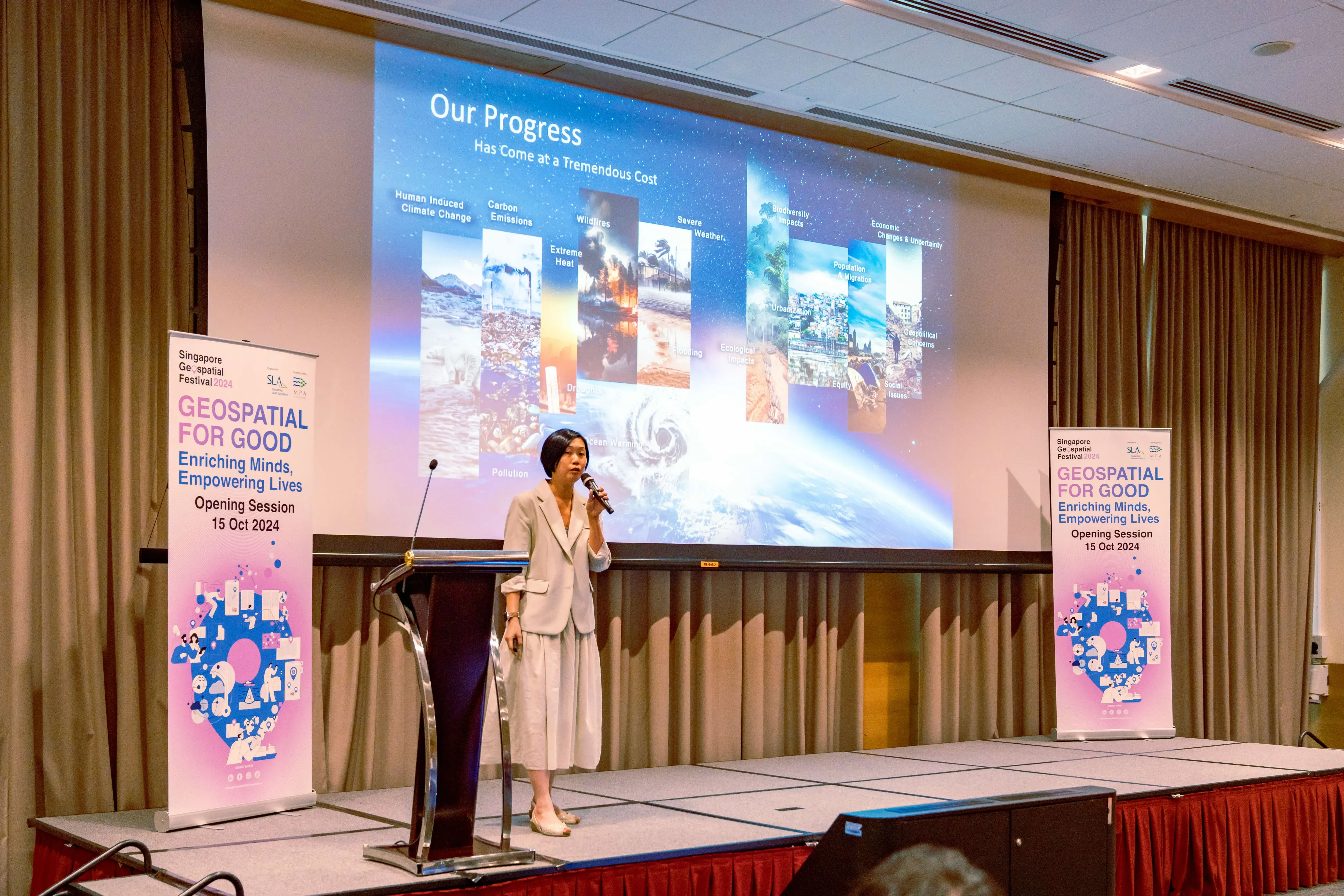 Woman in white outfit speaking on stage at Singapore Geospatial Festival 2024, with a slide titled "Our Progress" behind her.